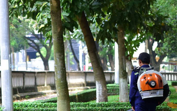 A gardener wearing a blower walks along a lush, tree-lined path in a city park.